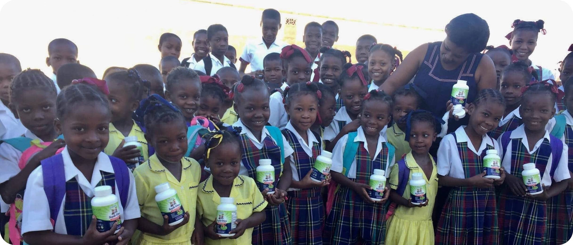 A group of smiling children in school uniforms holding bottles, gathered around an adult in a sunny outdoor setting.