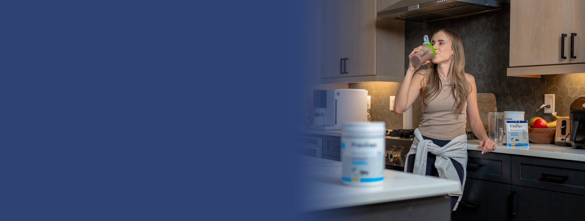 A woman in a kitchen drinking a Previnex shake, with Previnex products on the counter